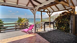 A covered outdoor area with a wooden deck, a pink chair, and a view of the sea. - Villa K'lia - Two Bedroom Cottage, Sleeps 4 (Le Robert)