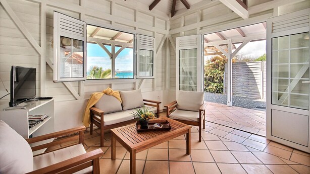 A spacious living area with a wooden coffee table, two sofas, and a TV. Large windows offer views of the outdoors. - Villa K'lia - Two Bedroom Cottage, Sleeps 4 (Le Robert)