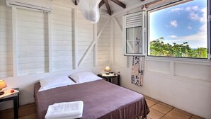 A bedroom with a bed, a nightstand, a lamp, a window with a view of greenery, and a ceiling fan. - Villa K'lia - Two Bedroom Cottage, Sleeps 4 (Le Robert)