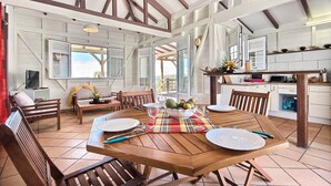 A spacious dining area with a wooden table set for four, surrounded by wooden chairs. A large window provides natural light, and the room features exposed wooden beams on the ceiling. - Villa K'lia - Two Bedroom Cottage, Sleeps 4 (Le Robert)