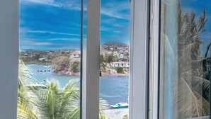 Vista costera a través de una ventana, con cielo despejado, palmeras y barcos en el agua.