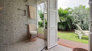 Un baño con ducha, inodoro y lavamanos. Hay una ventana con contraventanas blancas y vistas a un jardín con una silla y plantas.
