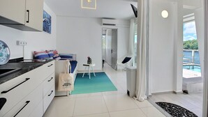 Een moderne keuken met witte kasten, een zwart aanrecht en een blauwgroen vloerkleed. Er staat een klein tafeltje met een vaas en een tas. Een witte stoel staat in de buurt van een glazen schuifdeur die uitkomt op een balkon met een zwembad.