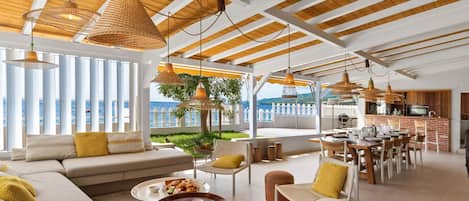 A spacious living area with a large sofa, dining table, and chairs, a kitchen area, and a view of the sea.