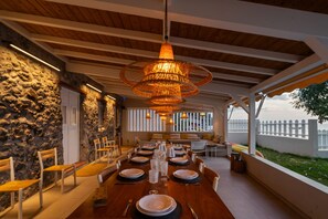 A dining area with a long wooden table set for a meal, surrounded by stone walls and a view of a white fence. - La maison dans la mer (Saint-Pierre)