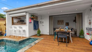 A poolside area with a wooden deck, a dining table with chairs, and a kitchen visible through an open door. - Villa Ti Pagerie - Two Bedroom Villa, Sleeps 4 (Les Trois-Îlets)