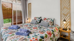 A bedroom with a floral bedspread, a wooden nightstand with a lamp, and a view of greenery outside. - Villa Ti Pagerie - Two Bedroom Villa, Sleeps 4 (Les Trois-Îlets)
