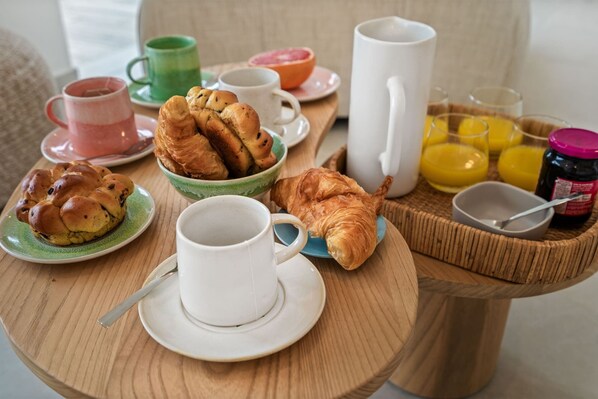 A breakfast table with croissants, a coffee cup, a juice pitcher, and a bowl of fruit.