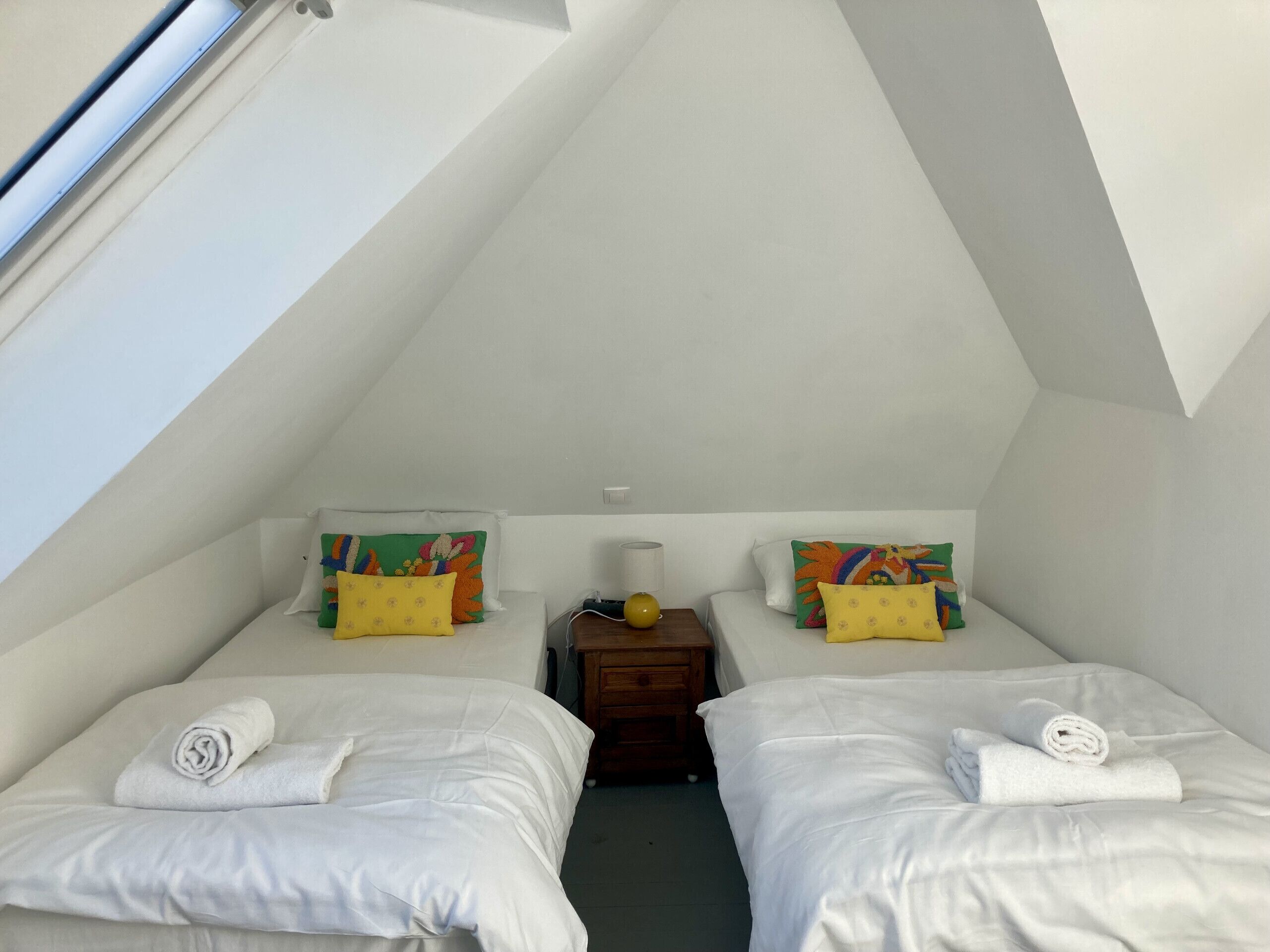 A small, triangular attic bedroom with two beds, each with a rolled towel, and colorful pillows.