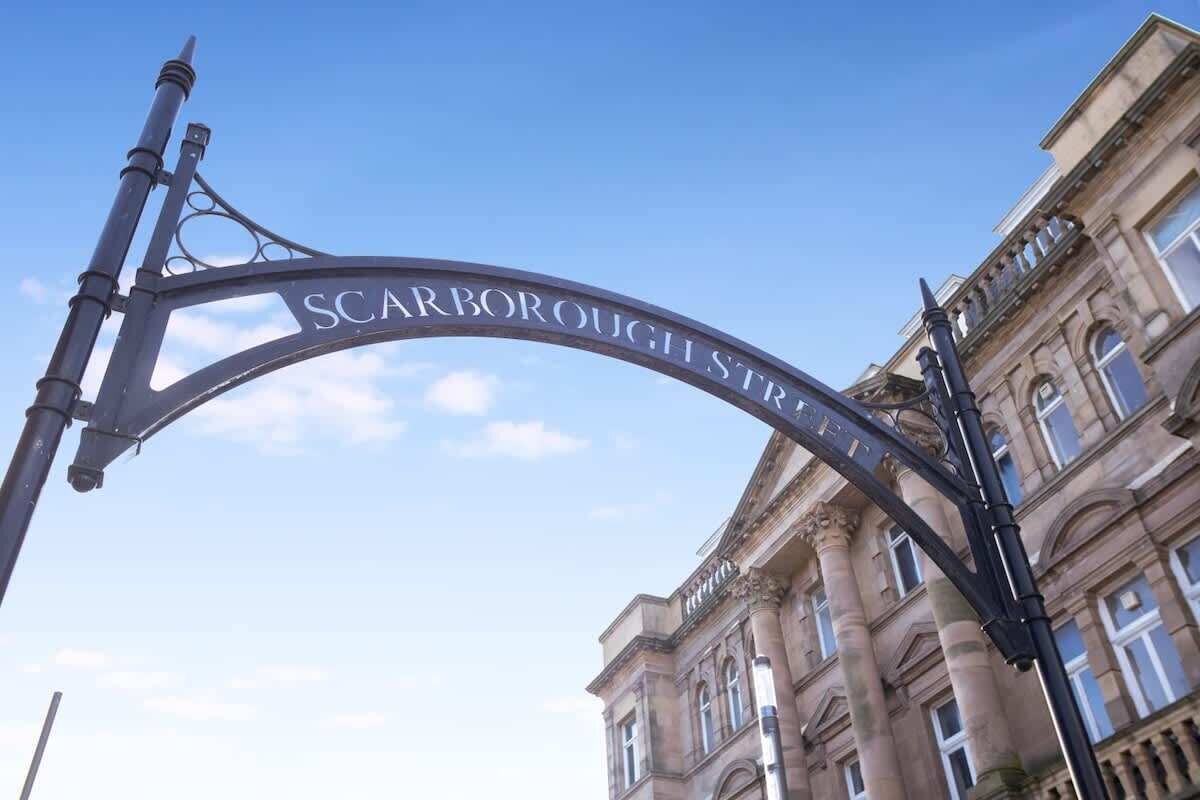 A metal sign with 'Scarborough Street' inscribed, mounted on a pole, with a classical building in the background.