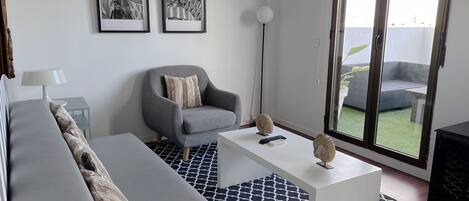 A modern living room with a grey sofa, a white coffee table, and a grey armchair. There are two framed pictures on the wall, a floor lamp, and a view of a balcony with a grey sofa.