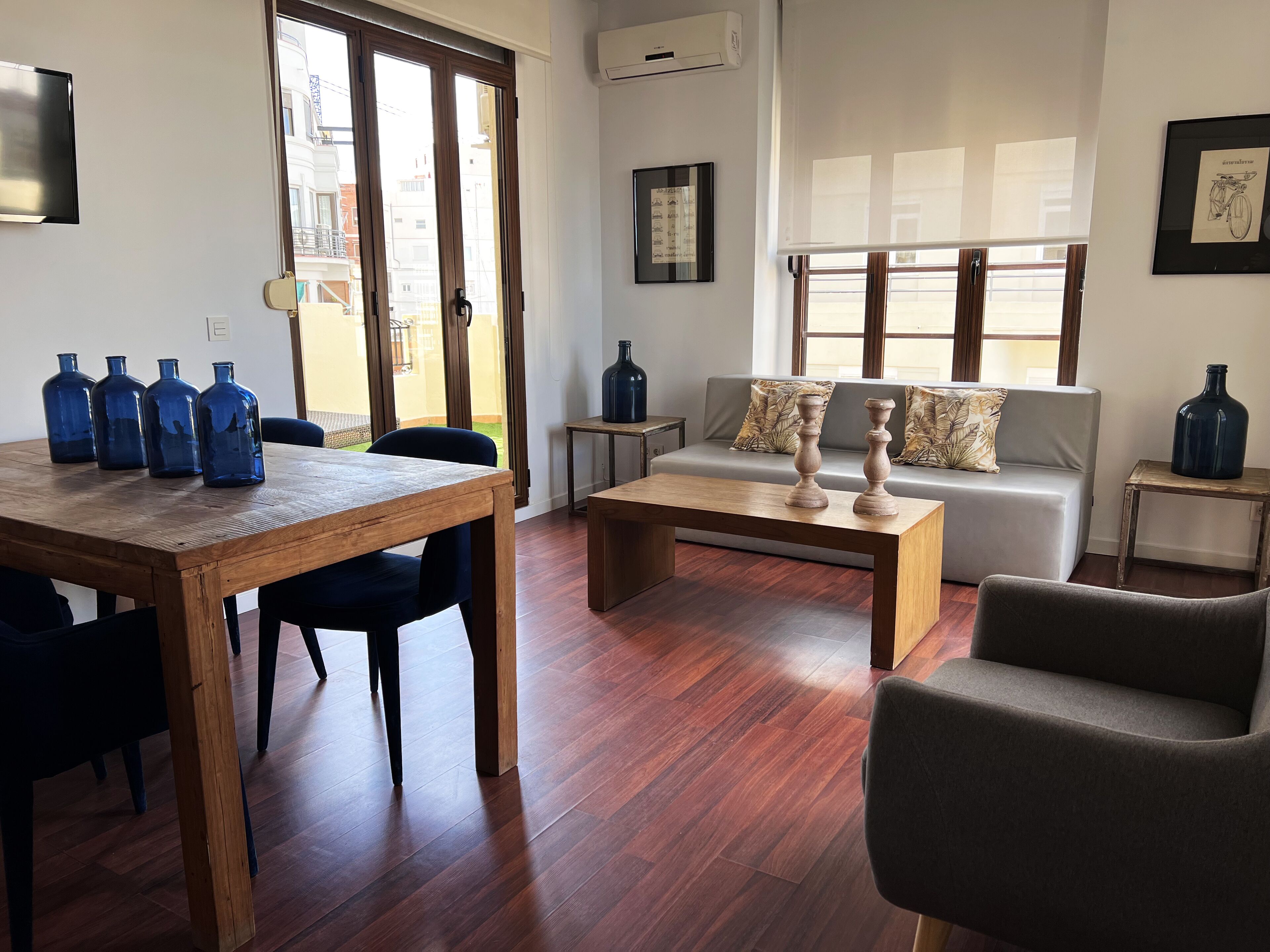 A living room with a wooden dining table, chairs, a sofa, and a coffee table. There are blue bottles on the table and a framed picture on the wall.
