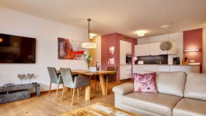 A modern living room with a wooden coffee table, a beige sofa with a deer pillow, a dining area with a wooden table and chairs, and a kitchen with white cabinets and a hanging light fixture.