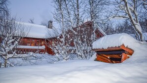 Una cabaña de madera con techo nevado, rodeada de árboles y una cerca de madera.