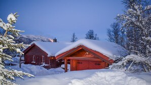 Una cabaña roja con garaje, rodeada de árboles cubiertos de nieve y una montaña al fondo.