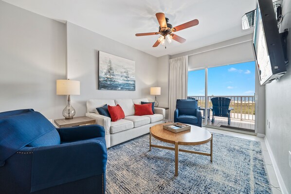 A modern hotel room with a white sofa, blue armchairs, a wooden coffee table, and a ceiling fan.