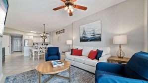 A modern living room with a white sofa, blue armchairs, a coffee table, and a ceiling fan. A kitchen is visible in the background with white cabinets and a dining area. - Tidewater 102- Orange Beach - 2bd/2bath - Beachfront (Orange Beach)