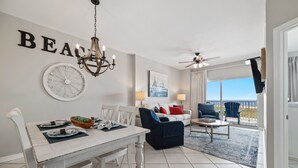A dining area with a white table and chairs, a chandelier, and a wall clock. There is a living area with a sofa and a coffee table, and a balcony with a view. - Tidewater 102- Orange Beach - 2bd/2bath - Beachfront (Orange Beach)