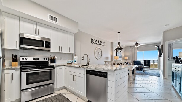A modern kitchen with white cabinetry, stainless steel appliances, and a 'BEACH' wall art. - Tidewater 102- Orange Beach - 2bd/2bath - Beachfront (Orange Beach)