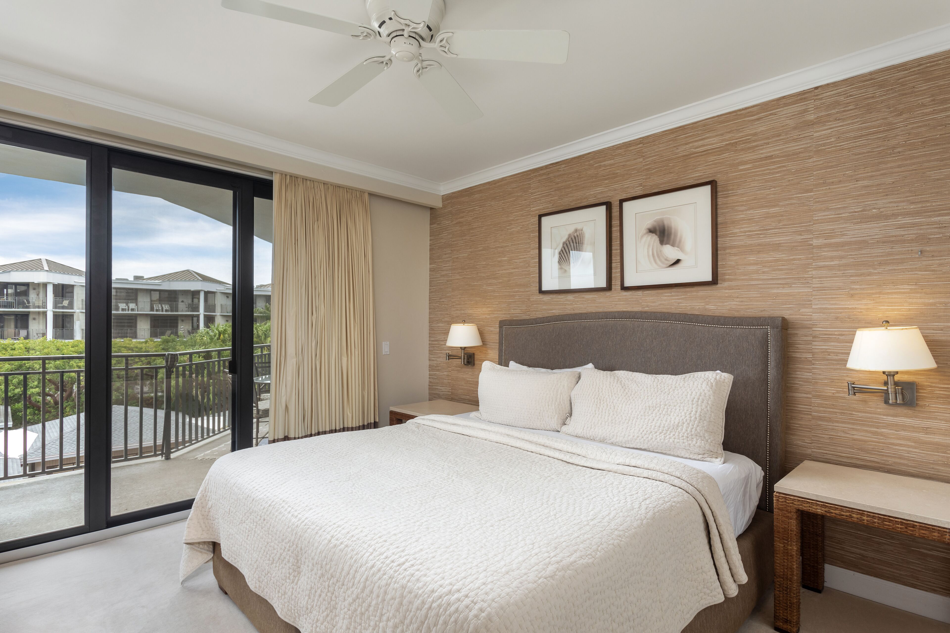 A hotel room with a large bed, a ceiling fan, a balcony with a view of other hotel buildings, and two framed pictures on the wall.