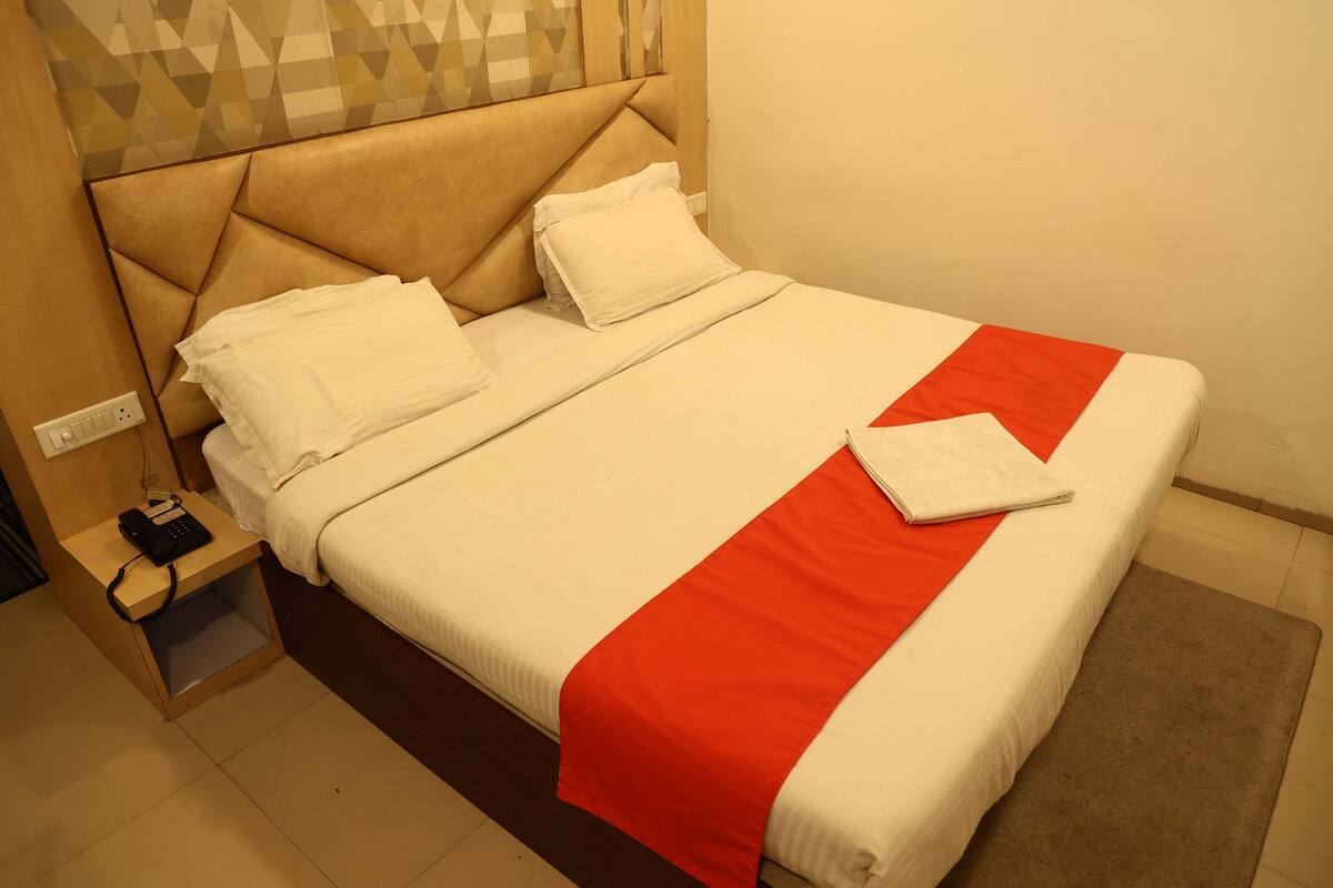 A neatly made bed with a red and white bedspread, a wooden nightstand with a phone and charger, and a wall with geometric patterned wallpaper.