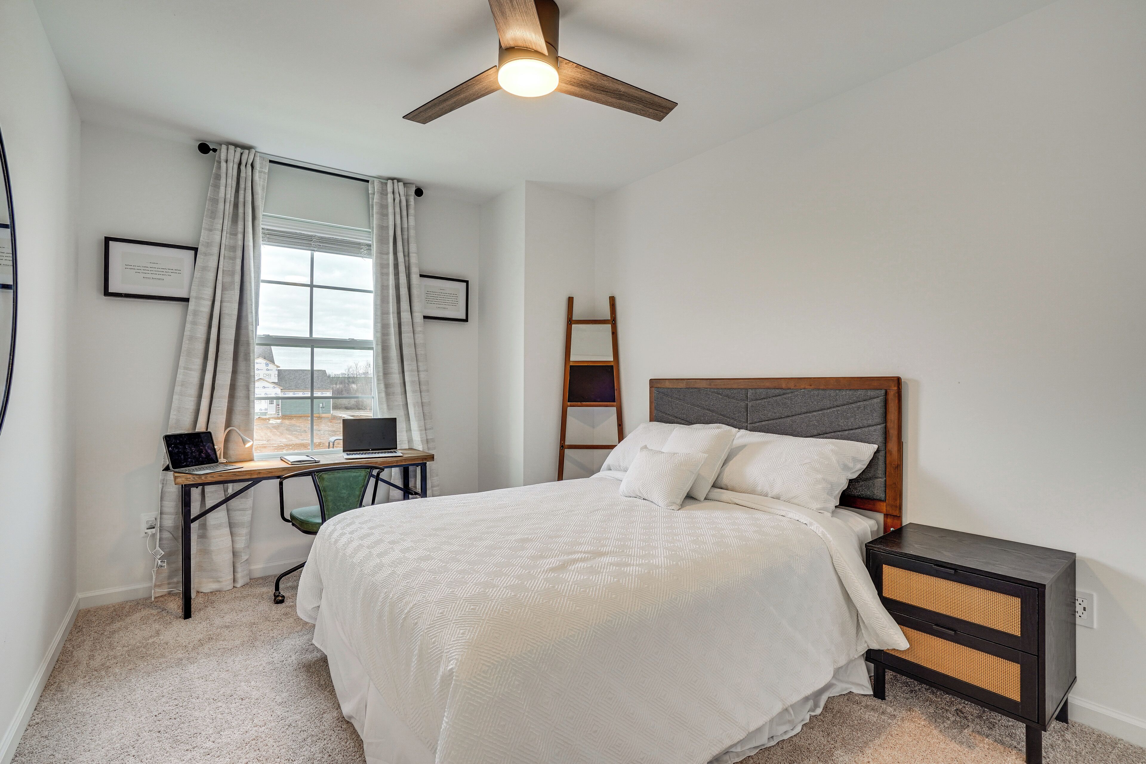 A bedroom with a bed, a desk with a laptop, a ceiling fan, and a window with curtains.