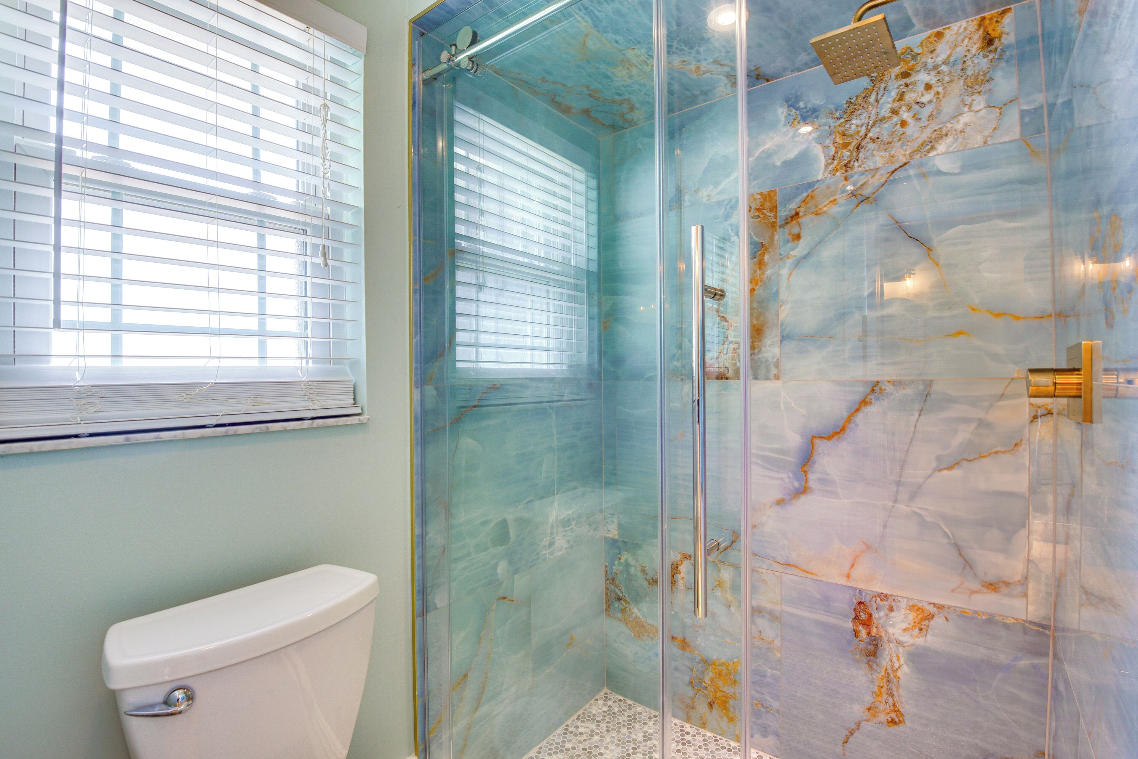 A bathroom with a glass-enclosed shower, a toilet, and a window with blinds.