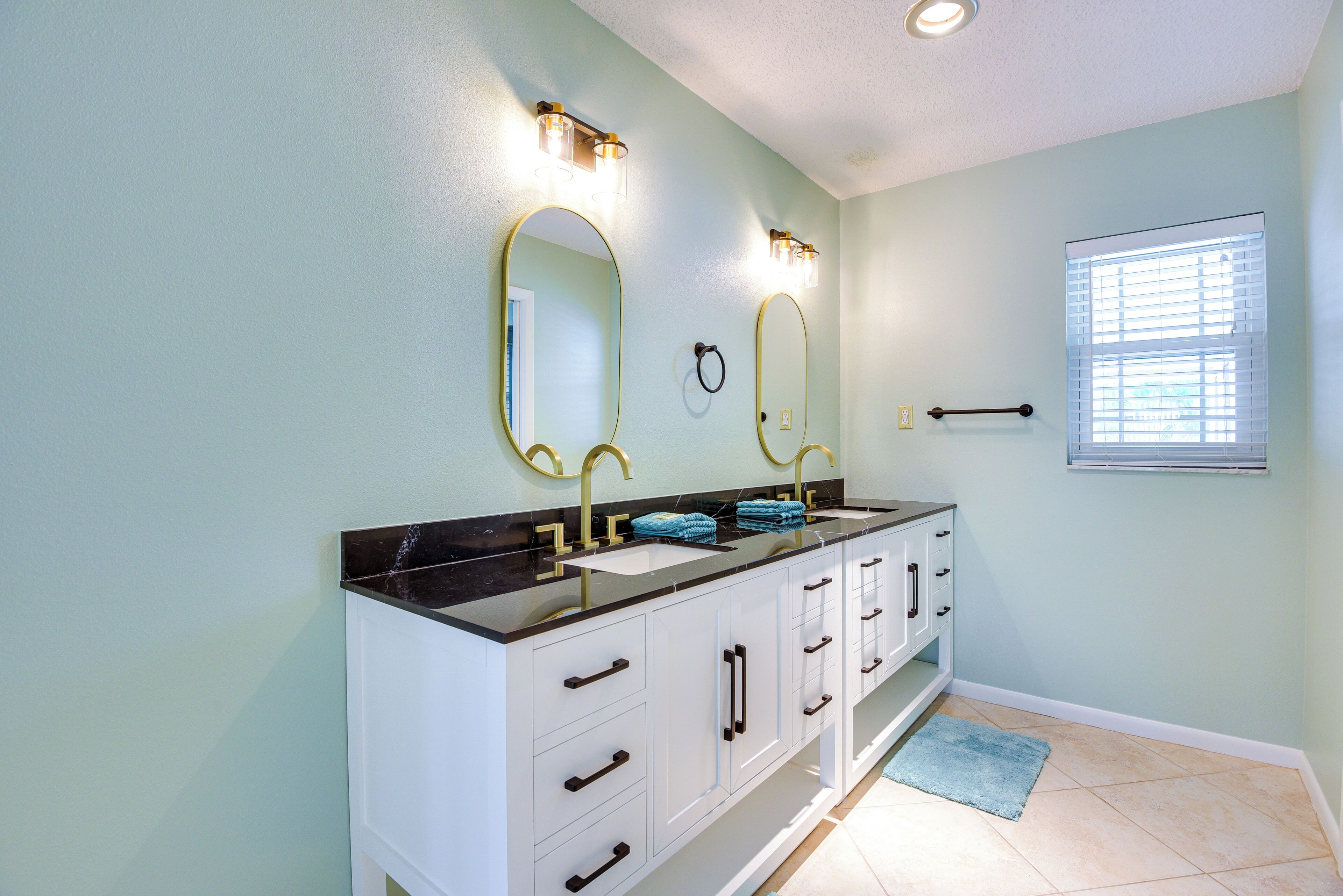 A bathroom with a double sink vanity, two wall-mounted mirrors, and a window with blinds.
