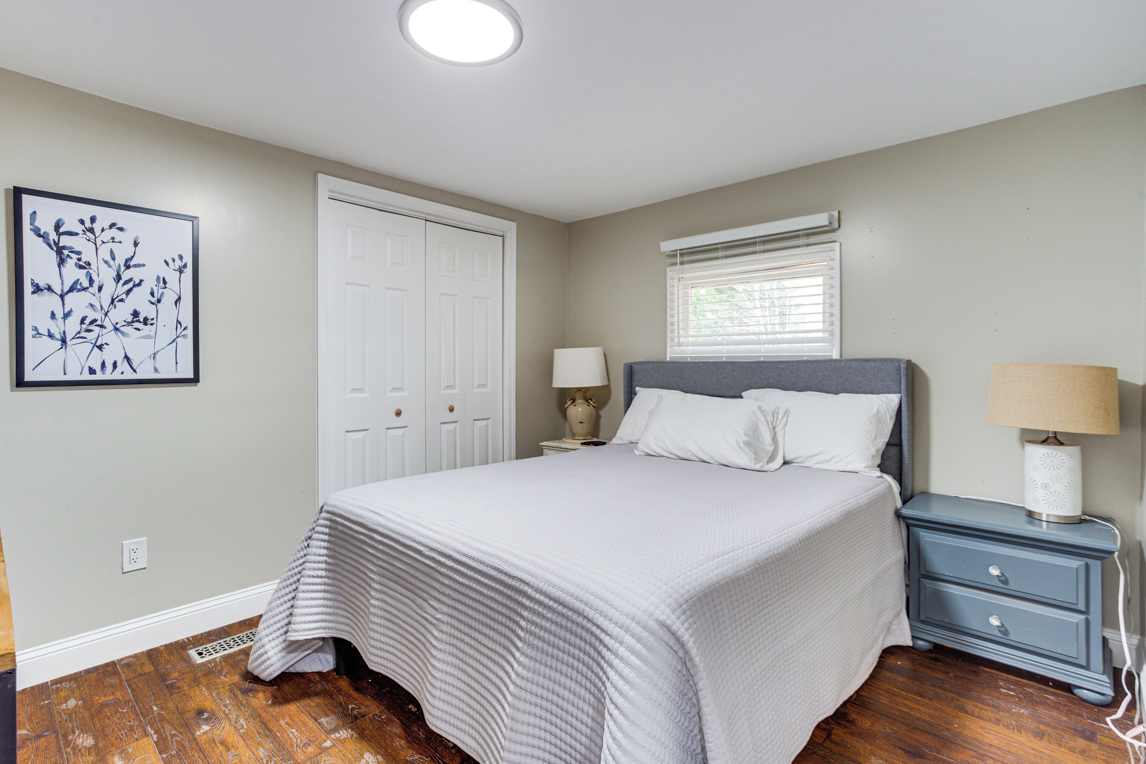 A bedroom with a bed, nightstands, a window with blinds, a door, and a wall-mounted light fixture.