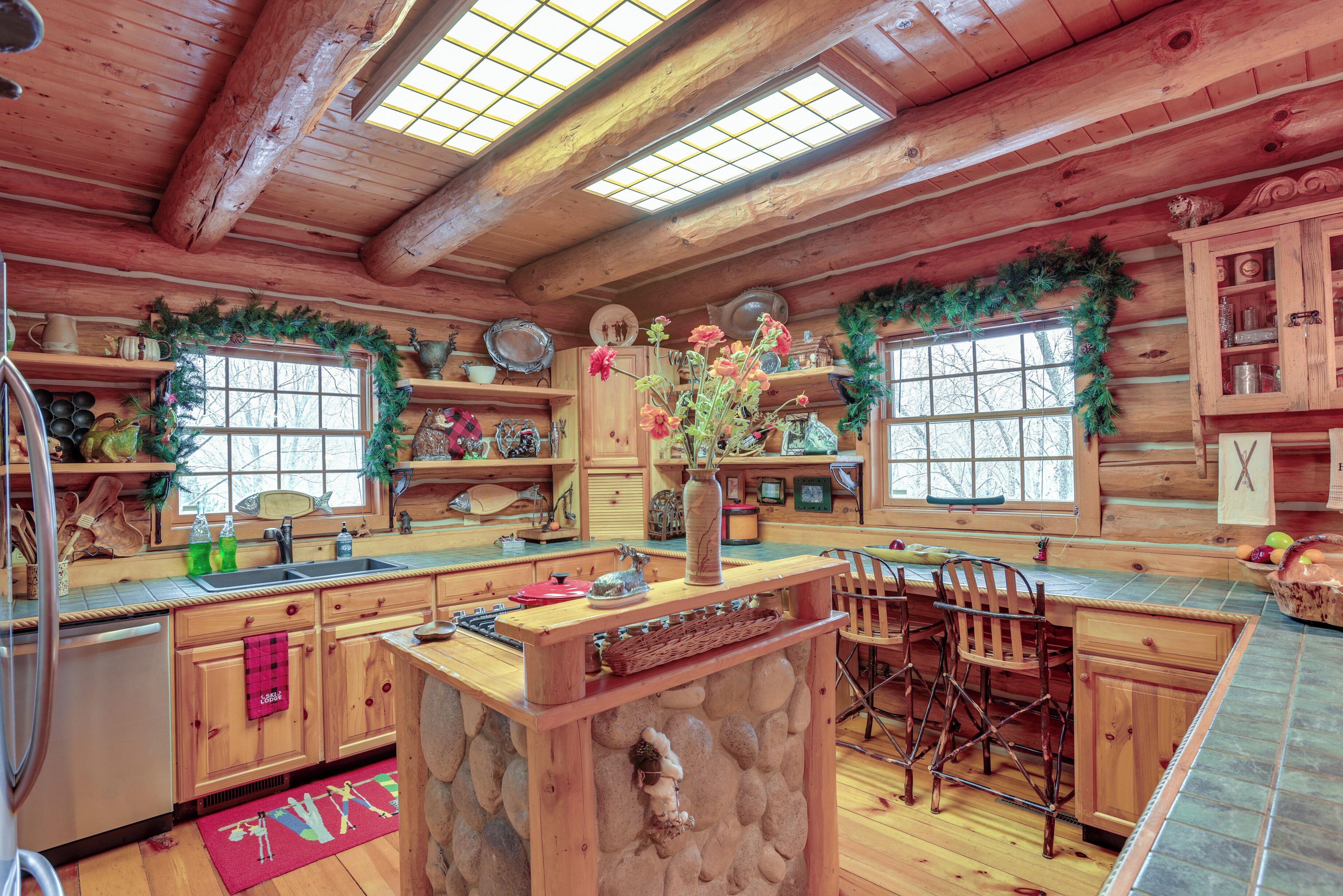 Een rustiek houten blokhutinterieur met een stenen kookeiland, houten kasten en een groot raam.