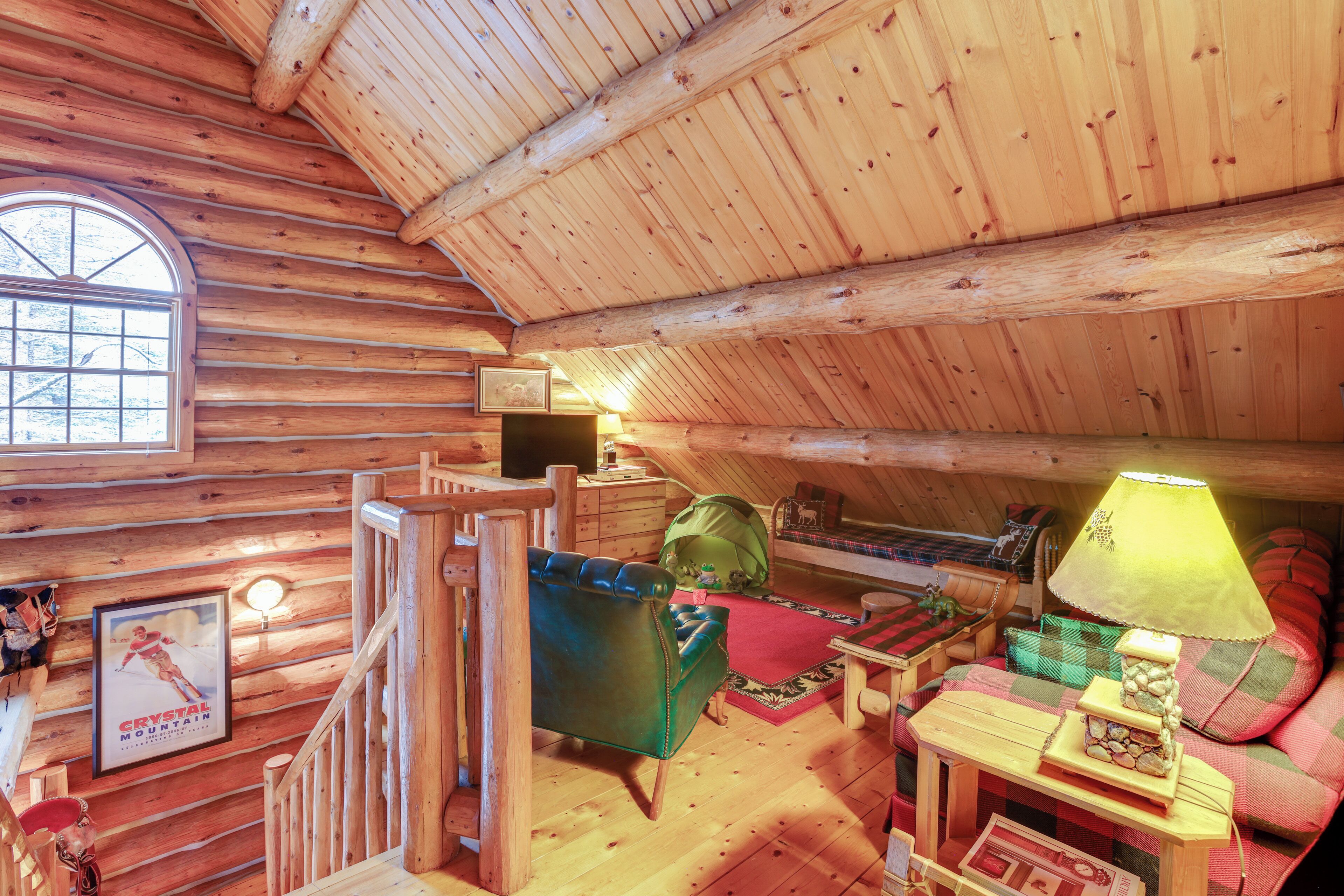 A log cabin interior with a wooden staircase, a green leather chair, a red and green patterned sofa, a lamp, and a framed picture on the wall.