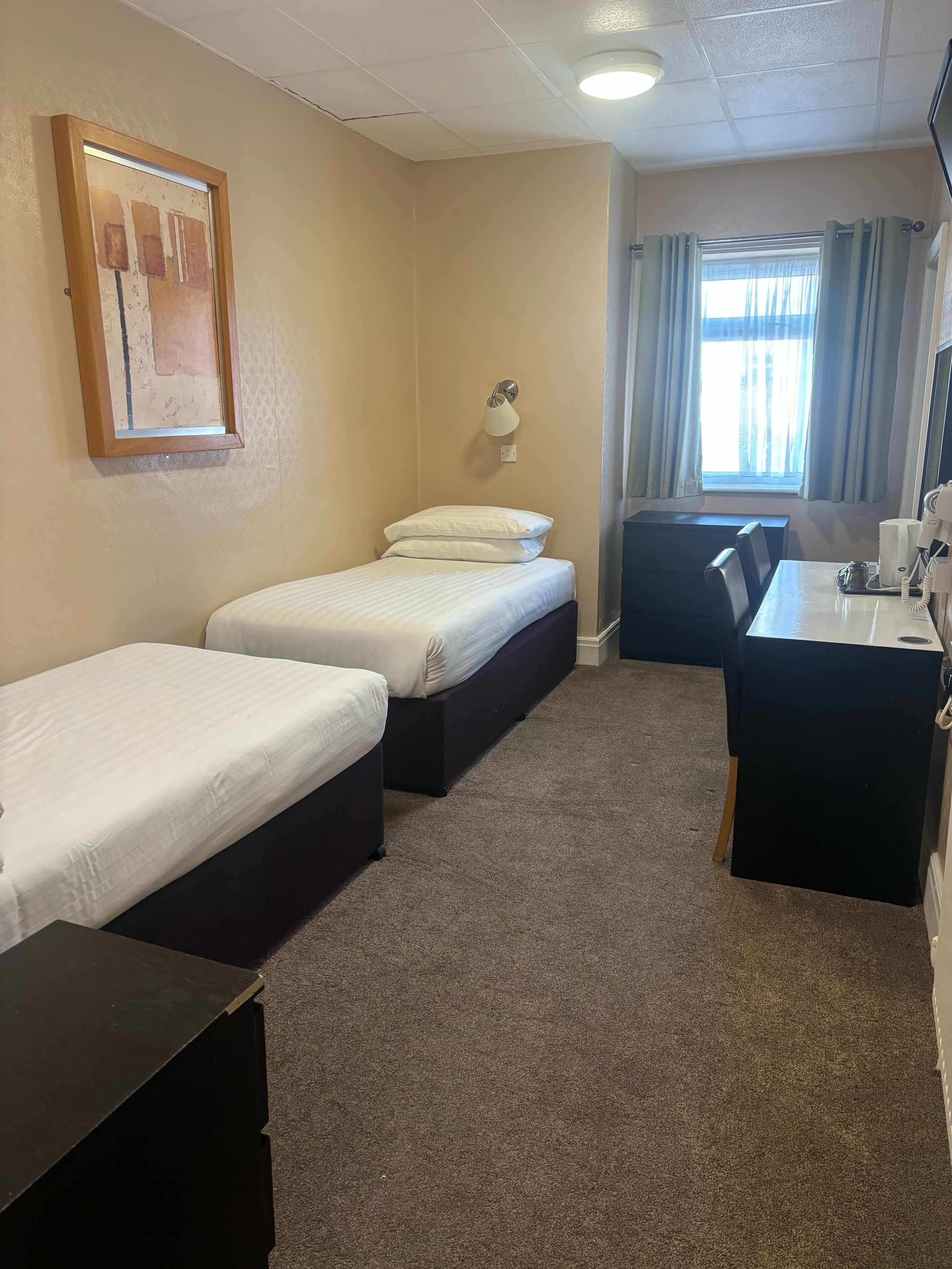 Habitación de hotel con dos camas individuales, un escritorio con teléfono, una lámpara y un ventanal con cortinas.