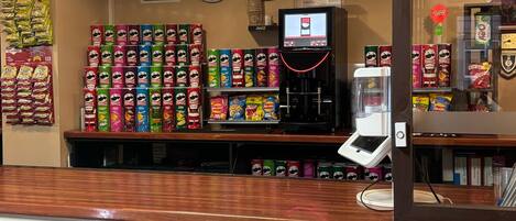 Een snackbar met diverse pakjes snacks en drankjes, een koffieapparaat en klokken aan de muur.