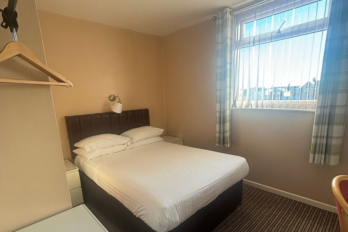 A hotel room with a bed, a bedside table, a hanging coat rack, a window with curtains, and a carpeted floor.