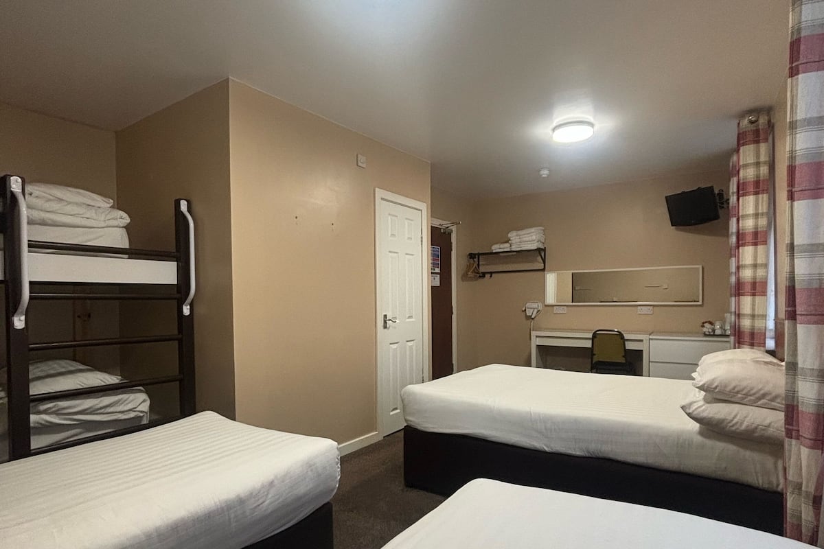 A hotel room with two single beds, a bunk bed, a desk with a chair, a mirror, and a television.
