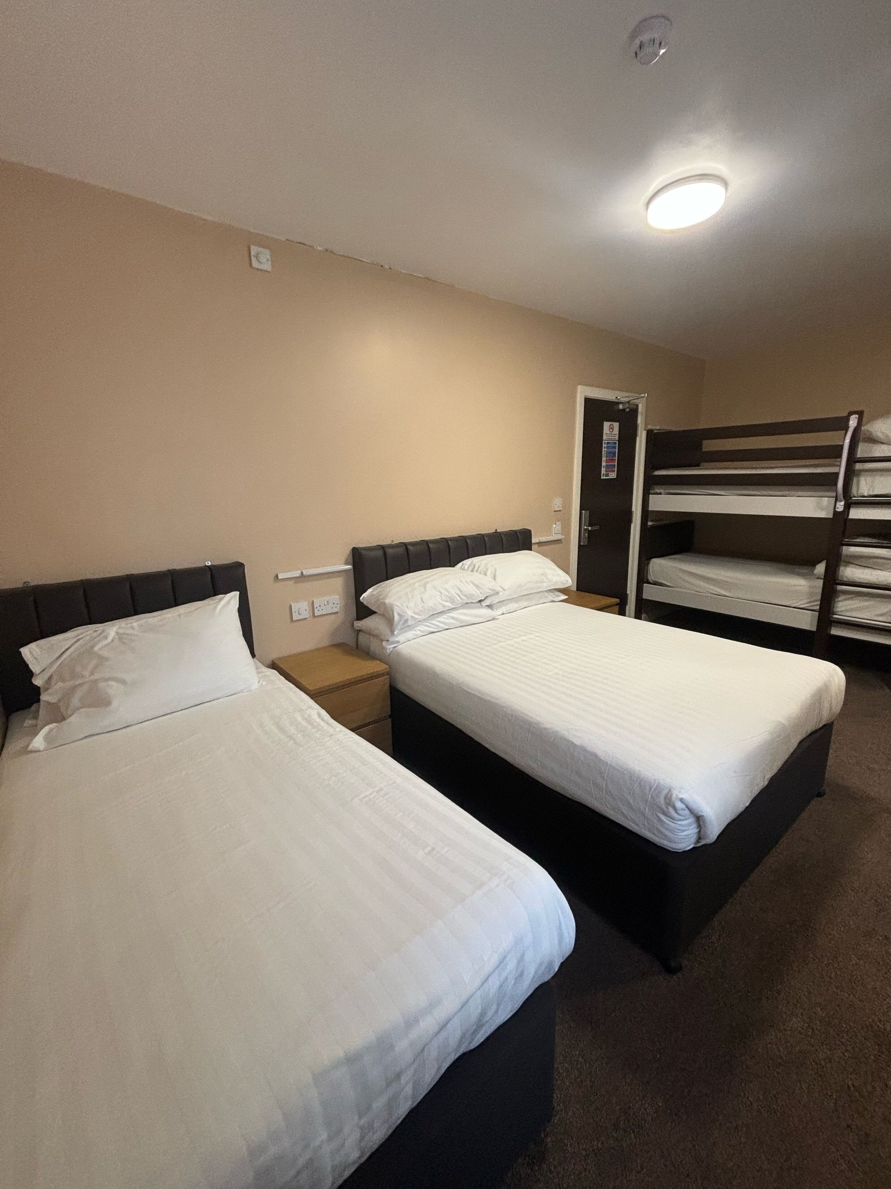A hotel room with two beds, a bunk bed, a door, and a ceiling light.