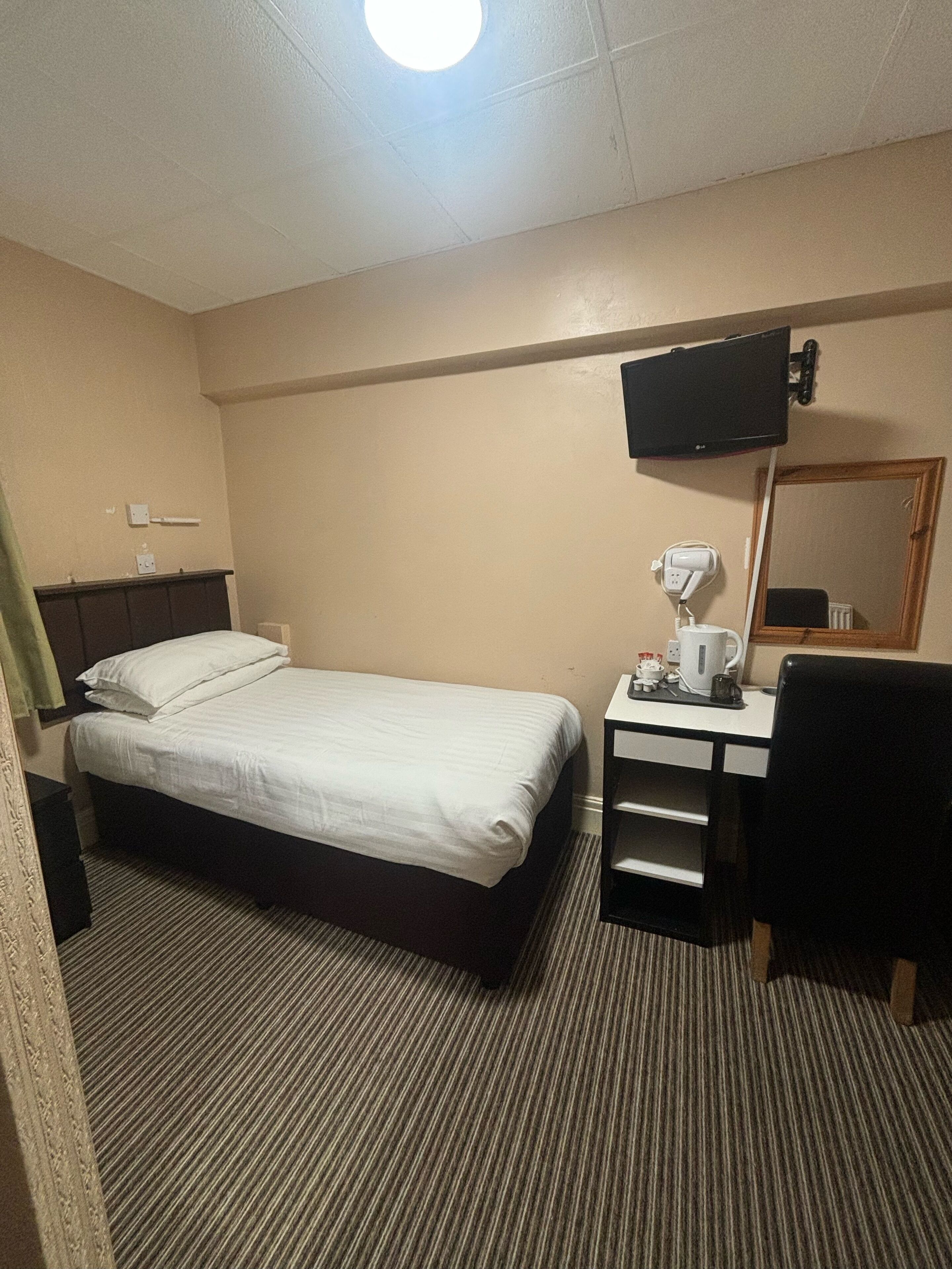 A hotel room with a single bed, a desk with a chair, a flat-screen TV mounted on the wall, and a mirror above the desk.