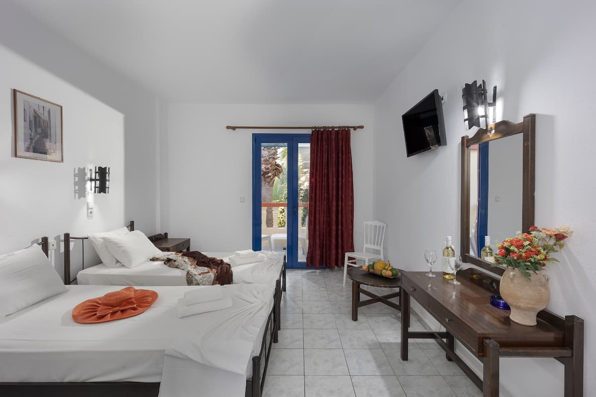 A hotel room with two beds, a wooden desk, a chair, a table with a vase of flowers, a television mounted on the wall, and a door leading to an outdoor area.
