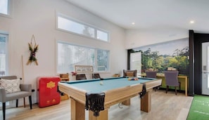 Una mesa de billar en una habitación con ventanas amplias, una silla roja y vistas a árboles a través del ventanal.