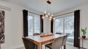 A dining area with a wooden table, chairs, a chandelier, and large windows with curtains.