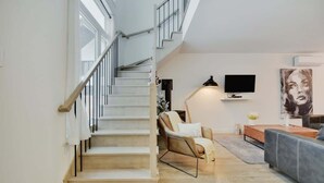 A staircase leading to an upper floor, a flat-screen TV mounted on the wall, a sofa, and a chair with a throw blanket.