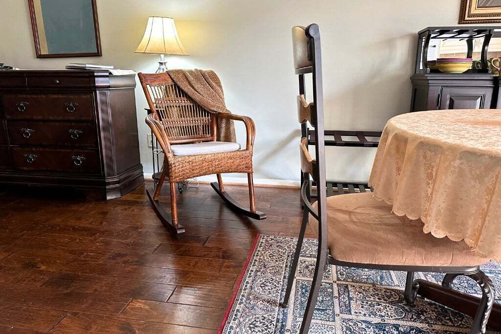 A room with a wooden dresser, a wicker rocking chair, a metal chair, and a table with a lace tablecloth.