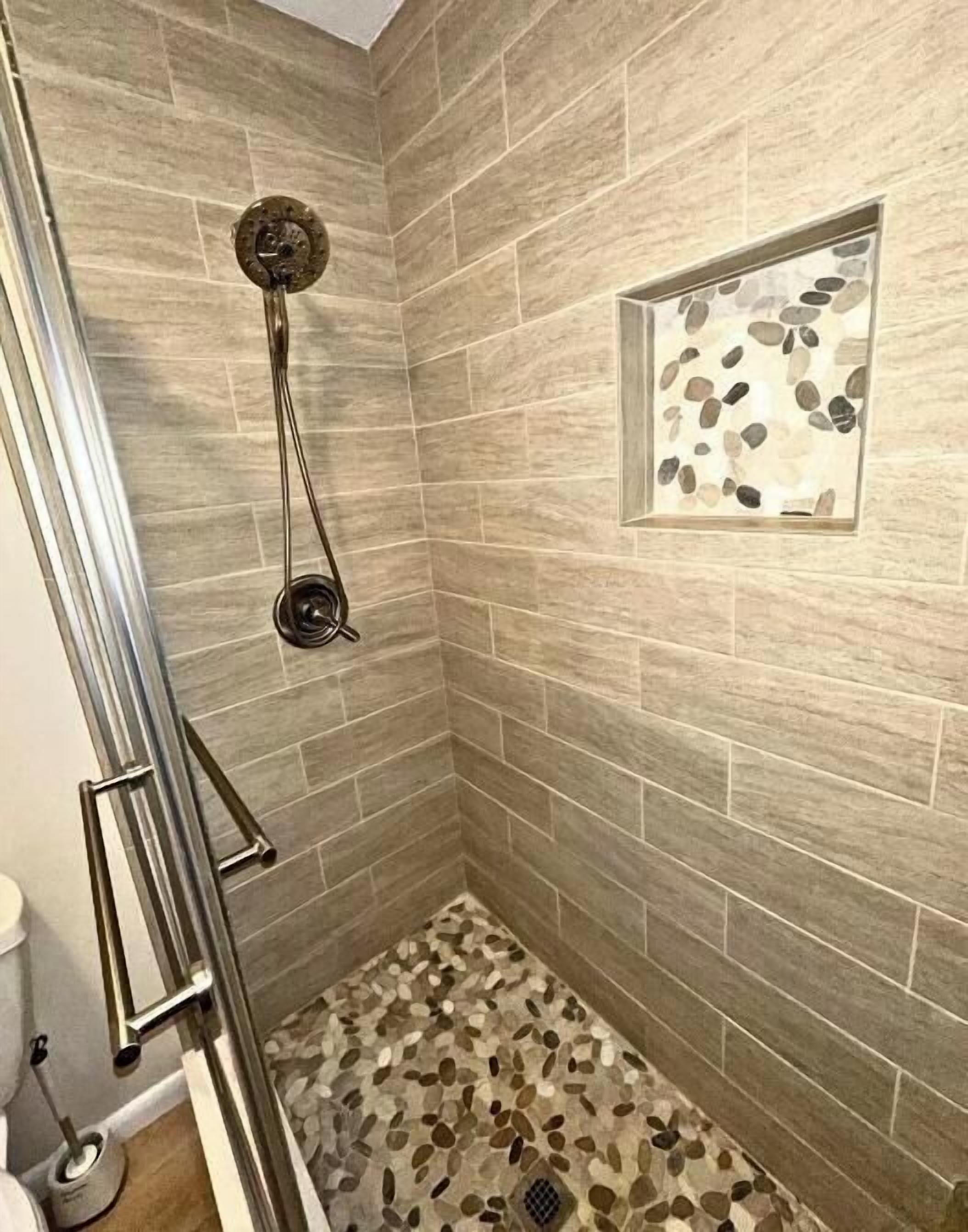 A shower with a glass enclosure, a metal handrail, and a pebble-patterned floor.