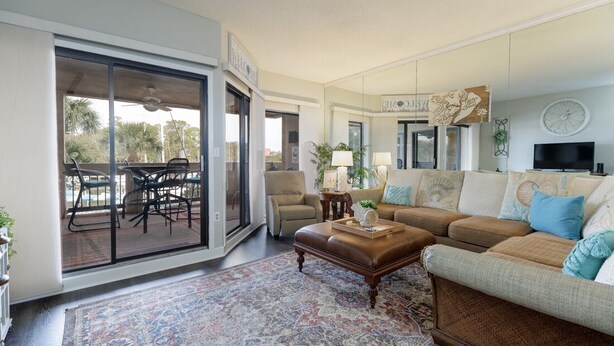 A spacious living room with a large sliding glass door leading to an outdoor patio area. There is a brown leather sofa, a patterned rug, and a coffee table with decorative items.