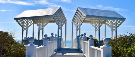 A wooden boardwalk leading to two white gazebos with lattice designs, surrounded by greenery and overlooking a body of water.