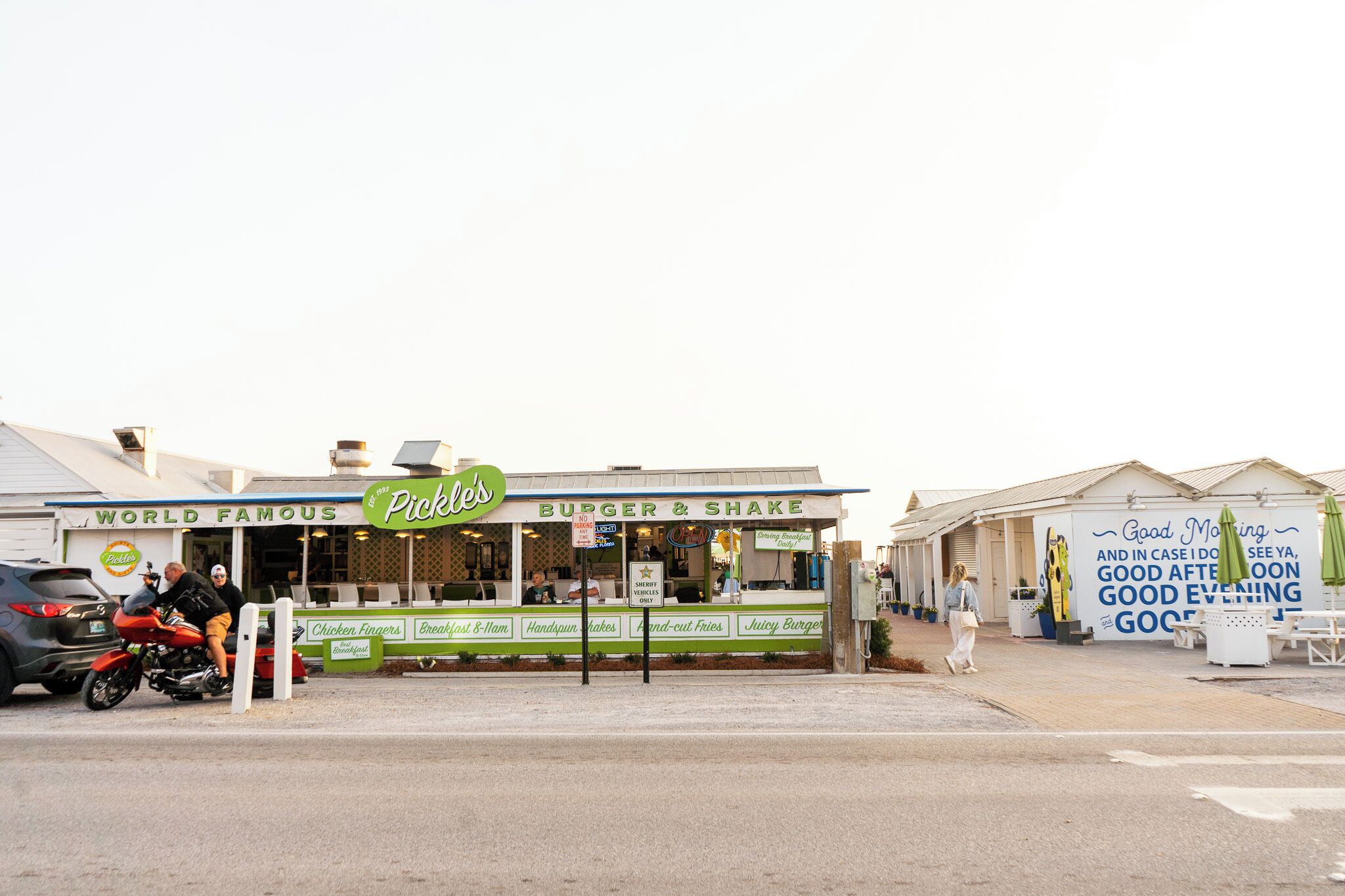 A roadside burger and shake stand with a green sign, a motorcycle parked in front, and a pedestrian walking by.