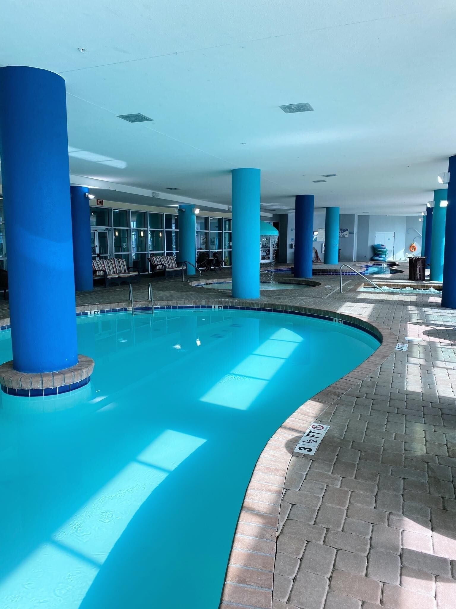 An indoor swimming pool with blue-tiled walls and columns, surrounded by seating areas and a tiled walkway.