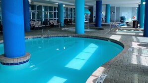 An indoor swimming pool with blue-tiled walls and columns, surrounded by seating areas and a tiled walkway. - 🌴Stunning Ocean Views | Pools + Hot Tub | Bay View Resort 1005🌴 (Myrtle Beach)