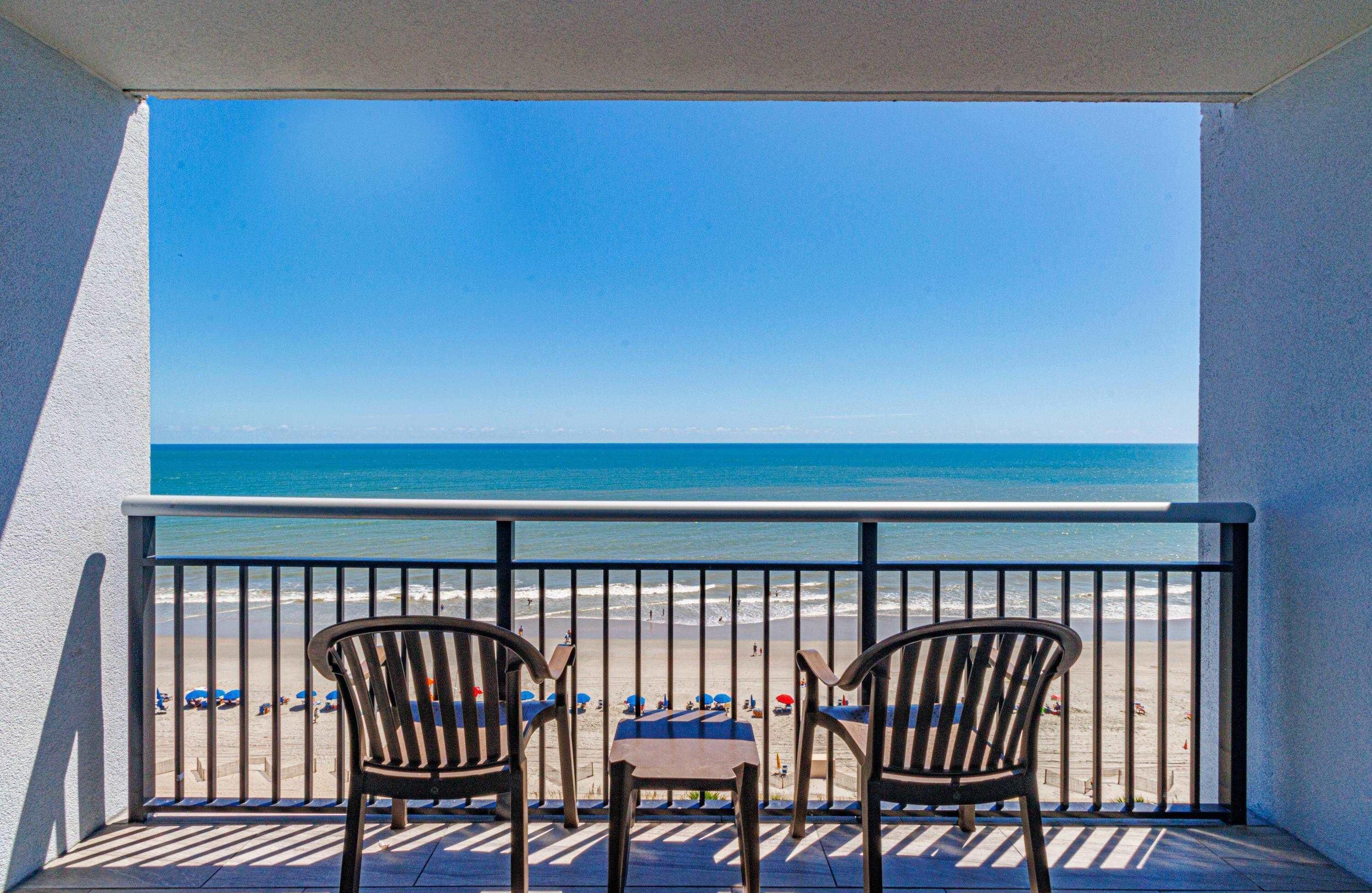 Un balcon avec deux chaises et une table donnant sur une plage parsemée de parasols et où se trouvent des gens.