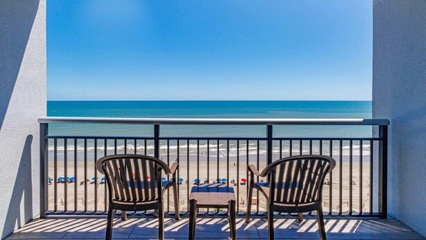 Varanda com duas cadeiras e uma mesa, com vista para uma praia com guarda-sóis e pessoas.